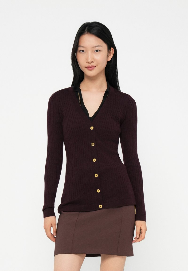 Young woman with straight black hair wearing a dark purple ribbed cardigan with gold buttons and a brown skirt, standing against a white background.