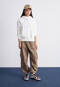 Haut à capuche blanc avec une poche avant et un logo, pantalon cargo beige avec poches latérales, baskets blanches et noires avec des accents marron, casquette beige.