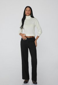 Crème coltrui met lange mouwen, gecombineerd met zwart hoge taille broeken en een bruine riem. Model staat voor een neutrale achtergrond.