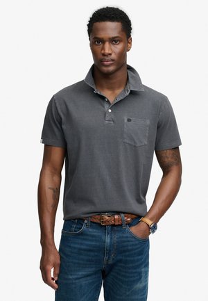 Homme aux cheveux courts et bouclés portant un polo à manches courtes gris foncé, un jean bleu, une ceinture tressée marron et une montre-bracelet, debout avec une main dans la poche.