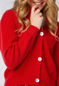 Czerwony kardigan prążkowany z dużymi, okrągłymi białymi guzikami i wysokim kołnierzem. Miękka tekstura z luźnym krojem, prezentujący pionowe linie w dzianinie.