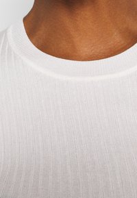 Weißes geripptes Strick-T-Shirt mit Rundhalsausschnitt, das eine glatte Textur und einen subtilen Glanz zeigt, mit feinen vertikalen Linien und einer taillierten Form.