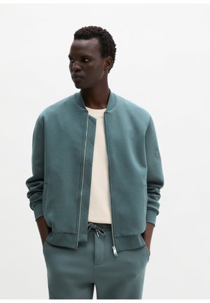 Jonge man met handen in zakken, gekleed in een teal zip-up jack en bijpassende broek over een wit shirt, kijkt naar links tegen een effen witte achtergrond.