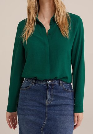 Vrouw draagt een groen lang mouwen overhemd met knopen, ingestopt in een blauw denim rokje met hoge taille, staand tegen een neutrale achtergrond.