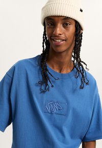 Camiseta de algodón azul con cuello redondo y mangas cortas; presenta un logo bordado de Vans en un azul más oscuro. El modelo lleva un gorro de punto blanco.