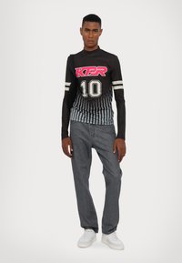 Cămașă neagră cu mânecă lungă, având logo-ul "KPR" și numărul "10", cu dungi verticale, asortată cu pantaloni gri și adidași albi. Design simplu.