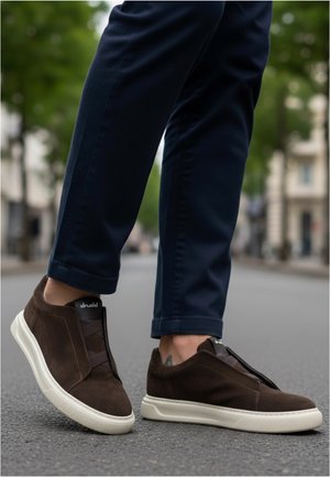 Person steht im Freien und trägt dunkelbraune Slip-On-Sneaker mit weißen Sohlen und marineblaue Hosen auf einer Asphaltstraße.