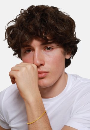 Giovane uomo con capelli ricci castani, che indossa una maglietta bianca e un braccialetto giallo, poggiando il mento sulla mano su uno sfondo neutro.