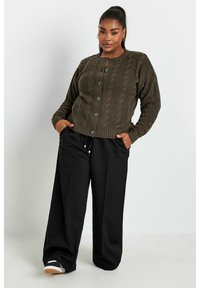 Olivgrüner, kabelgestrickter Cardigan mit rundem Ausschnitt und vorderem Knopfverschluss, kombiniert mit weiten schwarzen Hosen und Turnschuhen.