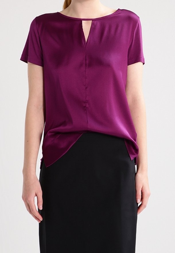 Femme portant un chemisier violet à manches courtes avec une petite fente à l'encolure devant et une jupe noire sur un fond uni.