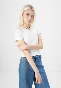 Fehér, rövid ujjú póló kerek nyakkal, magas derekú, világoskék farmernadrággal párosítva, amely laza szabású és látható varrással rendelkezik.