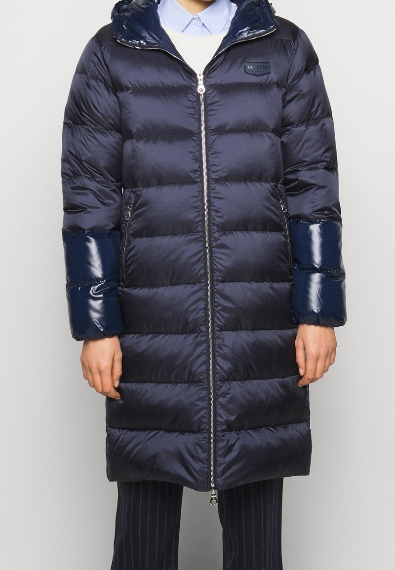 Marinblå pufferjacka med glänsande detaljer, dragkedja och horisontell quiltning. Har huva och ribbade muddarna. Lättviktig och isolerad design.