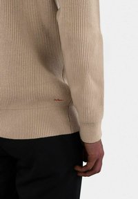 Beige ribbestrikket genser med lange ermer og en tilpasset nederkant, med en liten rød logo detalj nær bunnen. Glatt tekstur, uformelt design.