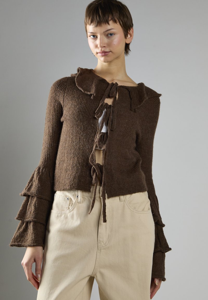 Brauner gestrickter Cardigan mit gerüschtem Kragen und schichtartig geschnittenen Ärmeln, versehen mit einem gebundenen Frontverschluss. Hellfarbene, enganliegende Hosen ergänzen das Outfit.