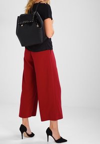 Mochila de cuero negro con un acabado texturizado y acentos de tachuelas. Combinada con pantalones de pierna ancha burdeos y tacones altos negros.