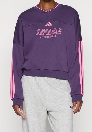 Osoba ubrana w fioletową bluzę Adidas z różowymi paskami na rękawach i różowym logo, w połączeniu z jasnoszarymi dresami Adidas.