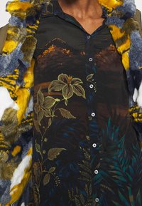 Camisa de botones con patrones florales y de follaje en tonos oscuros. Presenta un cuello de piel sintética en tonos de gris, amarillo y azul.