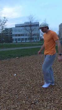 Mand i orange skjorte og grå bukser går på træflis i en park med moderne bygninger og bare træer i baggrunden ved skumring.