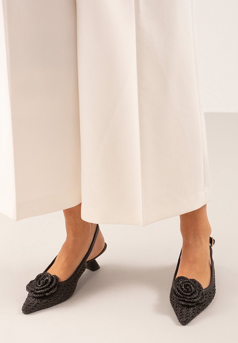Zapatos slingback negros con puntera afilada, fabricados en tela texturizada y con un adorno decorativo en forma de rosa en la puntera. Tacones bajos de bloque.