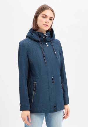 Frau trägt blaue Kapuzenjacke mit Reißverschluss und Knöpfen, Kordelzug an der Kapuze und Reißverschlusstaschen vorne, steht vor weißem Hintergrund.