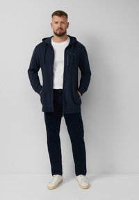 Navy zip-up hoodie met een voorzak, gedragen over een wit T-shirt, gecombineerd met donkere broek en witte sneakers. Glad textuur.