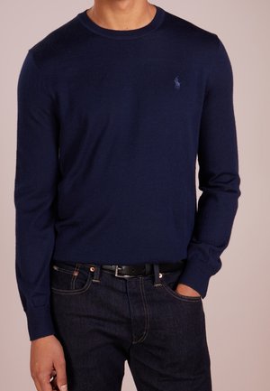 Mann trägt einen marineblauen langärmeligen Strickpullover mit kleinem gesticktem Logo auf der Brust, dunkle Jeans und schwarzen Gürtel, Hand in der Tasche.
