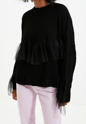 Schwarzer gestrickter Pullover mit transparenten Rüschendetails auf Brust und Ärmeln, getragen mit hellrosa Hose an einer Person.