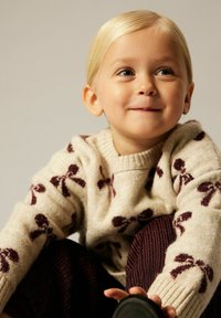 Pull en tricot beige avec des motifs de nœuds rouge foncé, encolure ronde et poignets côtelés, associé à un pantalon bourgogne texturé.