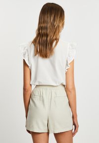 Weißes Blusenshirt mit Rüschensäumen aus Lochstickerei, kombiniert mit hellbeigen Shorts, die einen elastischen Bund und zwei Gesäßtaschen haben.