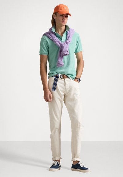 Mintgroene polo, paarse trui om de nek, crèmekleurige broek met versleten details en marineblauwe sneakers. Oranje pet en gestreepte riem.