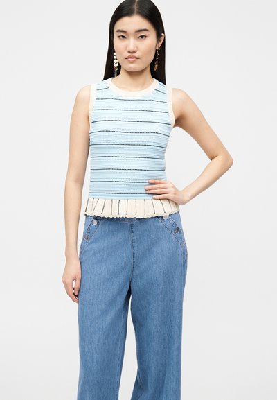 Γυναίκα με μακριά μαύρα μαλλιά που φοράει αμάνικο ριγέ τοπ με βολάν στο τελείωμα, σε συνδυασμό με ψηλόμεσα, φαρδιά μπλε τζιν παντελόνια.