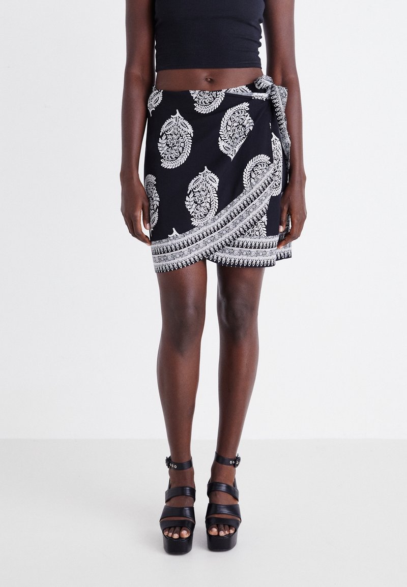 Femme portant une jupe portefeuille noire avec des motifs paisley blancs et des sandales compensées noires, vue de la taille jusqu'aux pieds.
