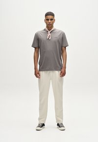 Szürke pamut rövid ujjú póló, kerek nyakkivágással és finom logó részlettel, világos krémszínű nadrággal és fekete tornacipővel párosítva. Vörös-fehér sál megkötve a nyak körül.