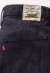 Jean en denim foncé avec un patch en cuir marqué, une étiquette rouge sur la poche et des détails de couture décorative. Design classique à cinq poches.