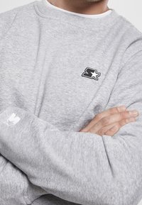 Sudadera gris hecha de material suave, con un logo en blanco y negro en el pecho y un pequeño logo en la manga. Diseño de cuello redondo.