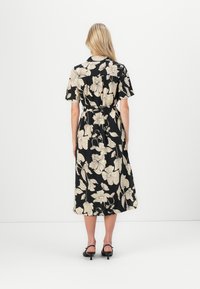 Robe midi noire avec un grand motif floral beige, manches courtes, taille ceinturée et ourlet fluide. Portée avec des sandales à talons noires.