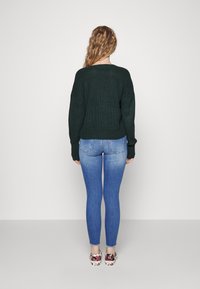 Pull en maille vert foncé avec épaules tombantes, associé à un jean skinny bleu clair et des chaussures à enfiler à motifs. Vue de dos.