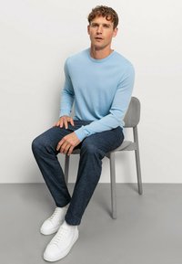 Hellblaustes Langarmshirt mit Rundhalsausschnitt, kombiniert mit dunkelblauen Jeans und weißen Sneakern. Sitzt auf einem grauen Stuhl vor einem hellen Hintergrund.