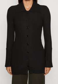 Svart, ribbad långärmad knapplöst cardigan med bred krage, lätt utsvängda manschetter, bärs över mörka ribbade byxor, framifrån.