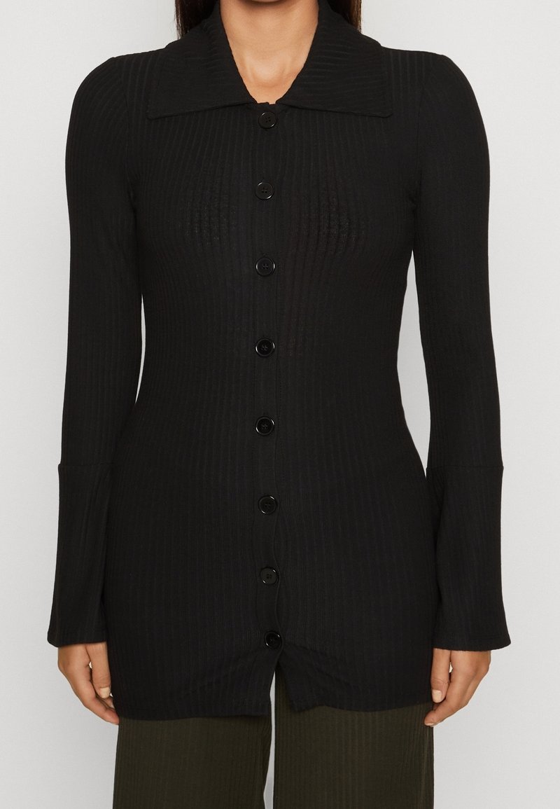 Svart, ribbad långärmad knapplöst cardigan med bred krage, lätt utsvängda manschetter, bärs över mörka ribbade byxor, framifrån.