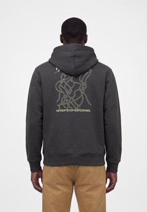 Mężczyzna w ciemnoszarej bluzie z kapturem z abstrakcyjnym wzorem linii i tekstem "Never Stop Exploring" na plecach, w połączeniu z jasnobrązowymi spodniami.