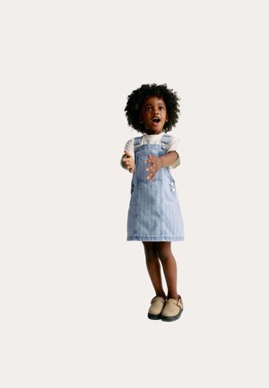 Vestido tipo peto de mezclilla con rayas azul claro, mangas cortas y botones en los bolsillos, combinado con zapatos beige tipo slip-on. Niño en una postura juguetona.