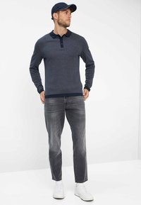 Polo a maniche lunghe blu navy con un motivo testurizzato e colletto scuro, abbinato a jeans grigi a gamba dritta e scarpe da ginnastica bianche.