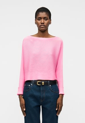 Femme portant un pull côtelé rose vif à encolure bateau, rentré dans un jean taille haute bleu foncé avec une ceinture noire.