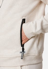 Felpa beige chiaro con una tasca con zip. La zip è nera con un tira-zipp in tessuto che presenta la scritta "DENIM FACTORY". Tessuto morbido e strutturato.