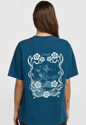 Woman with long brown hair wearing a teal O'Neill t-shirt featuring a floral frame and a figure diving in the center on the back.