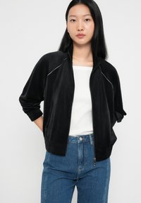 Chaqueta bomber de terciopelo negro con cremallera, que presenta un ribete blanco. Mangas largas, puños acanalados y un ajuste relajado. Combínala con jeans de tiro alto.