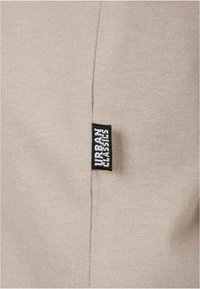 Beige stoff med fin strikketekstur, med en svart etikett som har teksten "URBAN CLASSICS" sydd på siden.