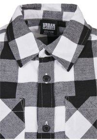 Camisa de cuadros en blanco y negro, con cuello puntiagudo, dos bolsillos frontales, botones negros y detalles de costura visibles. Etiqueta Urban Classics en el interior.