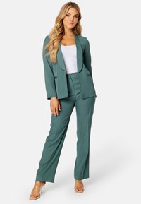Teal trouwpak met een getailleerde blazer met een notched lapel en op maat gemaakte broek. Glad textuur met een elegante silhouet, gecombineerd met heldere sandalen met hakken.